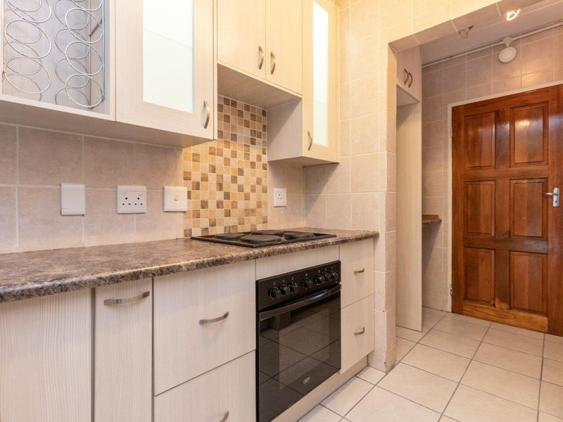 A functional kitchen featuring light-toned cabinetry, a tiled backsplash, built-in oven and hob, and tiled flooring, leading to a separate doorway.