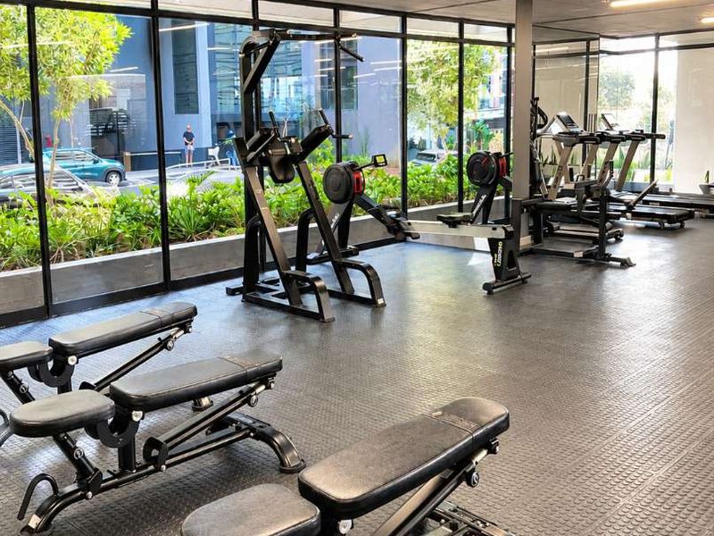 A modern, well-lit apartment complex gym featuring rubber flooring, floor-to-ceiling windows, and an exposed concrete ceiling.