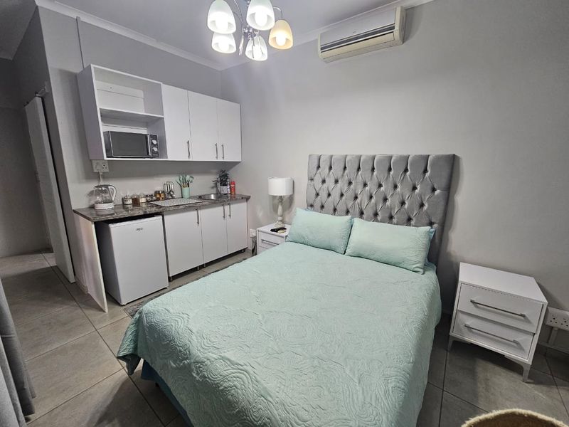 Compact open-plan studio apartment featuring a kitchenette with built-in cabinetry, tiled flooring, and a wall-mounted air conditioning unit.