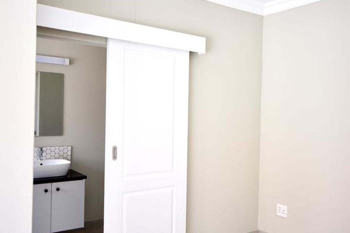 A clean, neutral bedroom featuring tiled floors, crown molding, and an en-suite bathroom with a modern vanity and sliding door access.