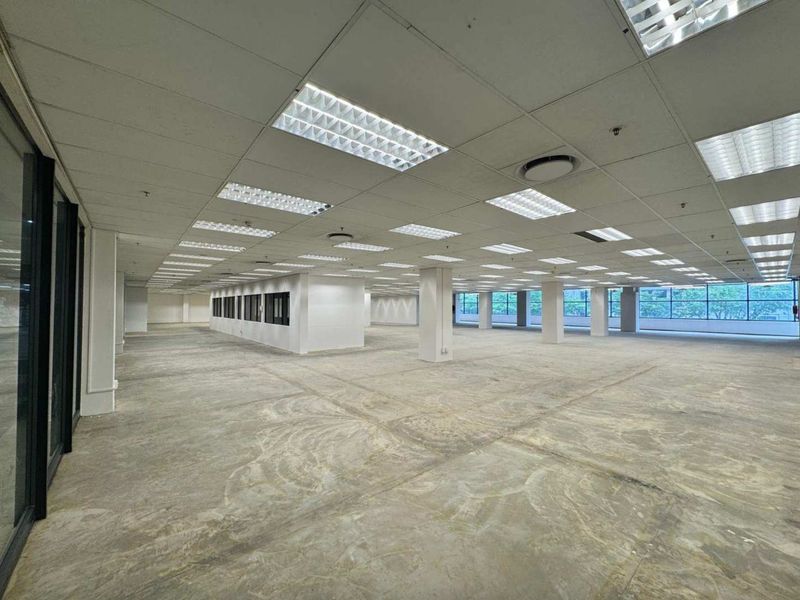 A large, open-plan commercial office space featuring a suspended ceiling, recessed lighting, structural columns, and expansive windows with unfinished concrete flooring.