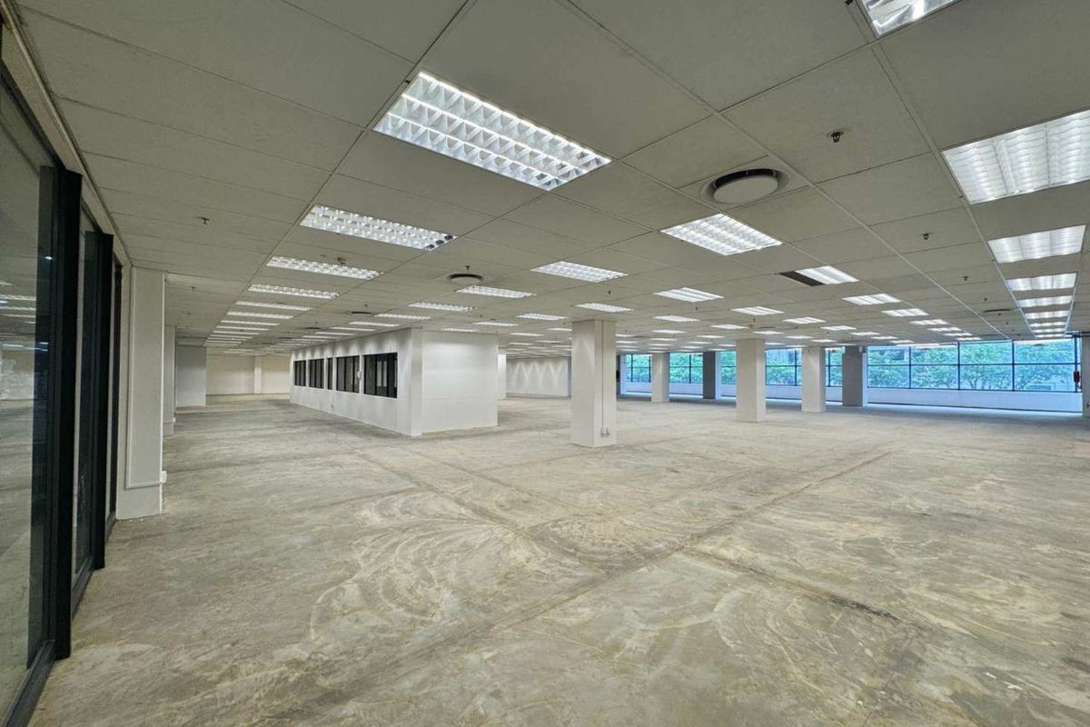 A large, open-plan commercial office space featuring a suspended ceiling, recessed lighting, structural columns, and expansive windows with unfinished concrete flooring.