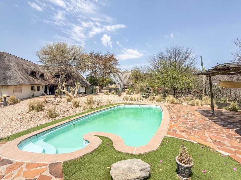 Outdoor pool area featuring a swimming pool, stone patio, and a pergola, set against a natural landscape with a thatched-roof dwelling in the background.