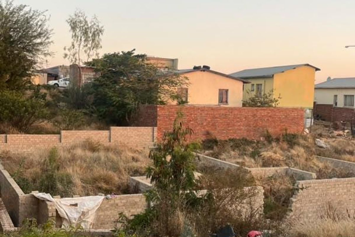 A vacant residential plot featuring unfinished brick foundation walls and boundary fencing, situated in a developed neighborhood setting.