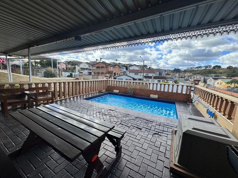 A covered outdoor patio area featuring a swimming pool with brick paving and decorative balustrades, offering an elevated view of the surrounding neighborhood.
