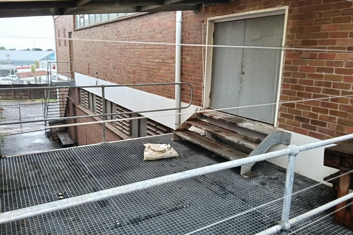 Industrial rear exterior area featuring a metal grating platform, safety railings, and a metal staircase leading to a double door entrance.