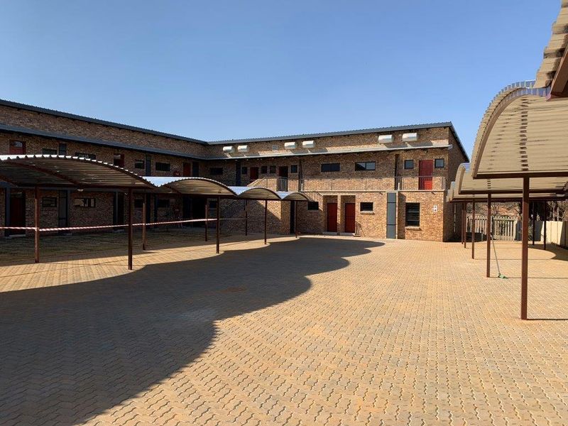 Exterior view of a multi-story apartment complex featuring a paved courtyard, covered parking carports, and exposed brick facade.