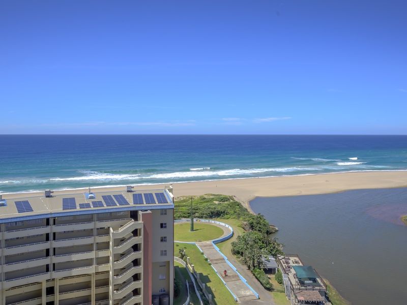 Elevated coastal view overlooking the ocean, beach, and estuary, featuring a multi-story apartment building with rooftop solar panels.
