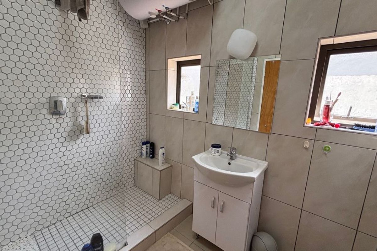 A functional bathroom featuring a walk-in shower with hexagonal tiling, a vanity basin, and a wall-mounted electric water heater.