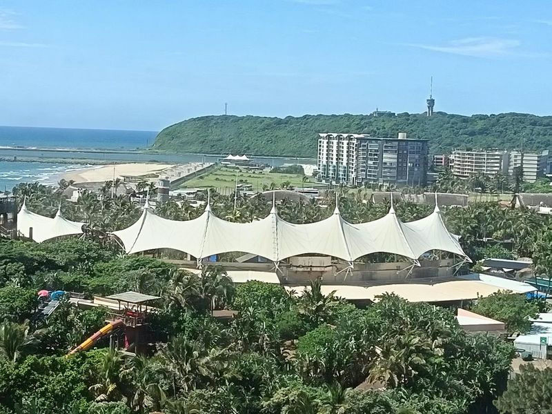 Elevated panoramic view overlooking the coastal landscape, featuring a large tensile structure, lush greenery, and the ocean in the distance.