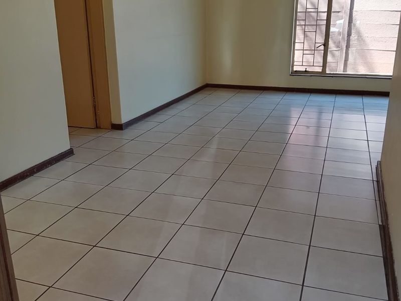 A bright, empty living area featuring neutral ceramic floor tiles, clean walls, and a large window with security bars.