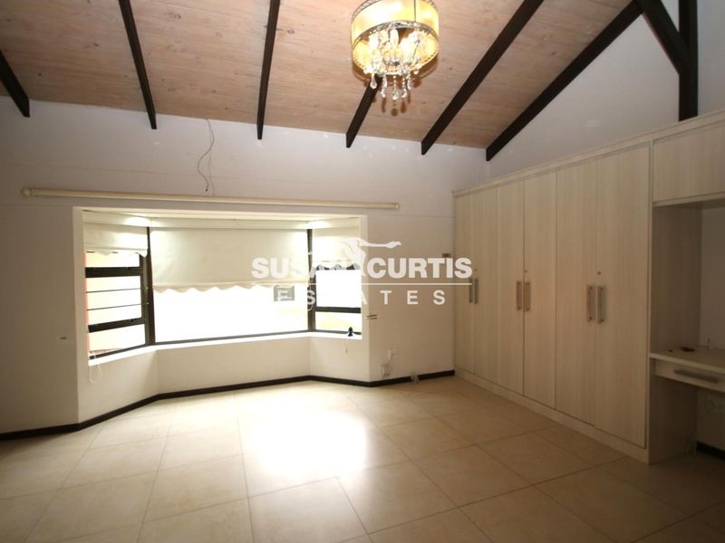 Spacious bedroom featuring a vaulted timber ceiling with exposed beams, built-in cupboards, an integrated desk, and a large bay window providing natural light.