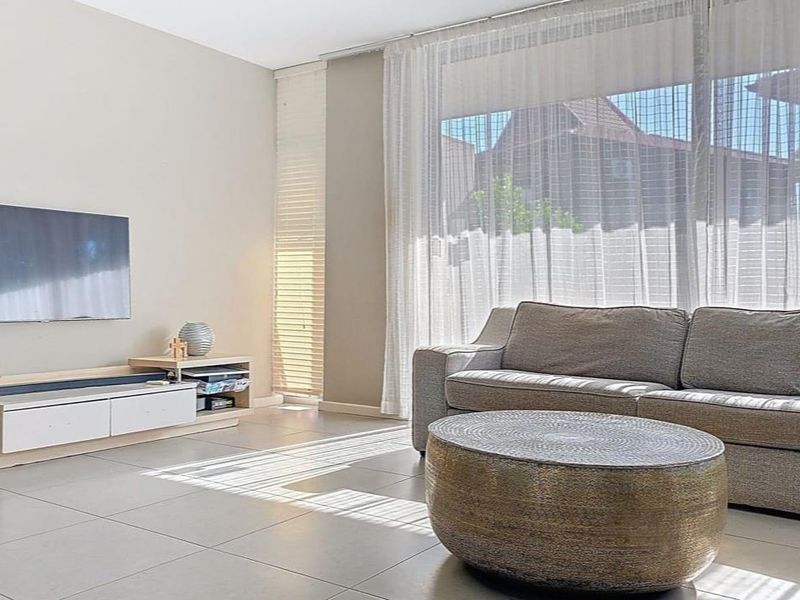 A bright and modern living room featuring large windows with sheer curtains, venetian blinds, and neutral tiled flooring.