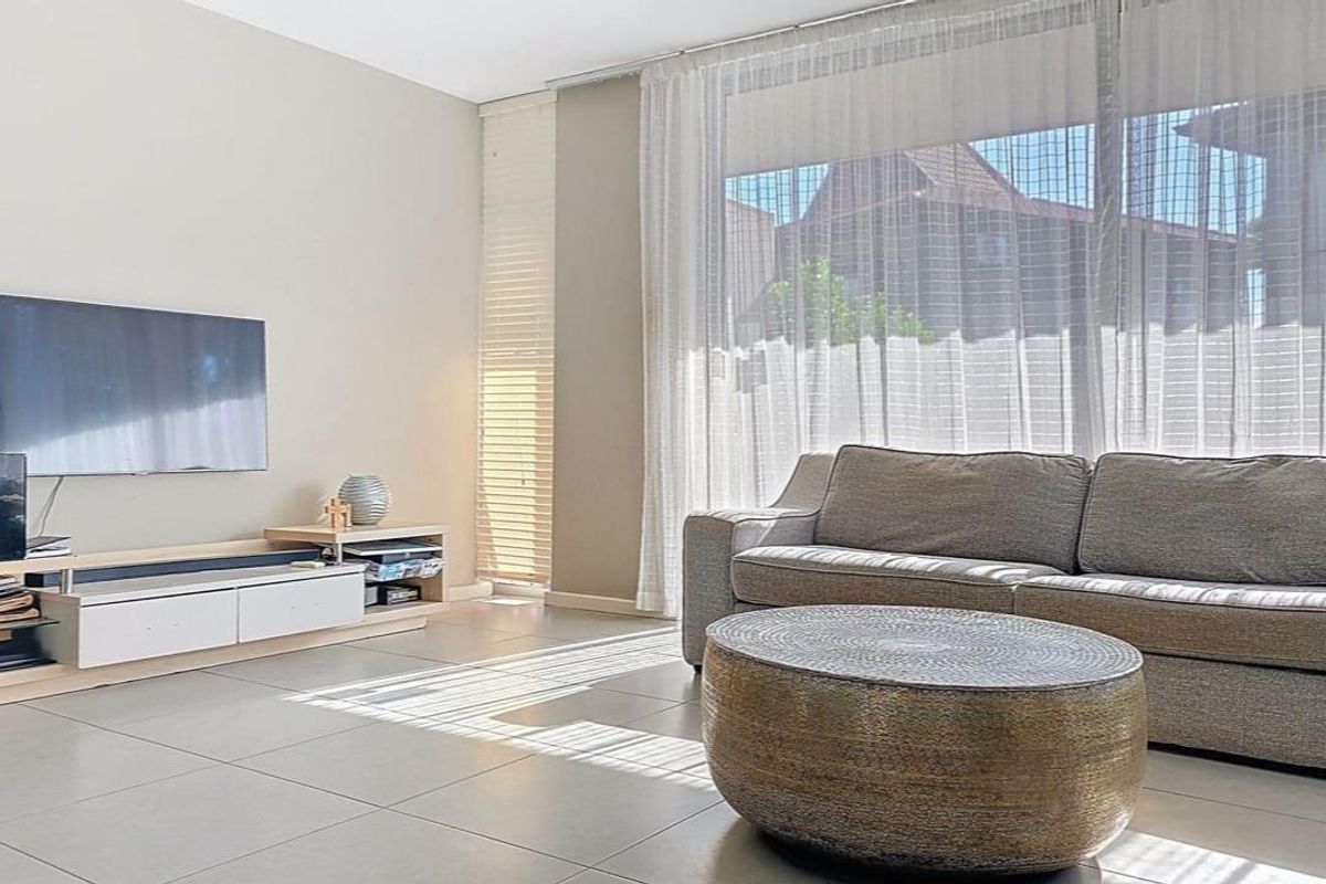 A bright and modern living room featuring large windows with sheer curtains, venetian blinds, and neutral tiled flooring.