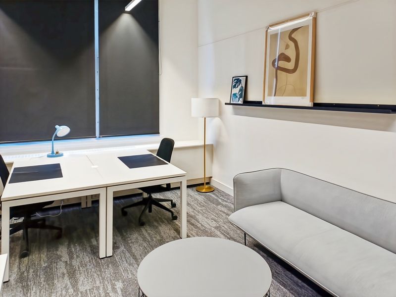 A modern, well-lit office space featuring neutral tones, carpeted flooring, roller blinds, and a clean, professional layout suitable for a productive work environment.