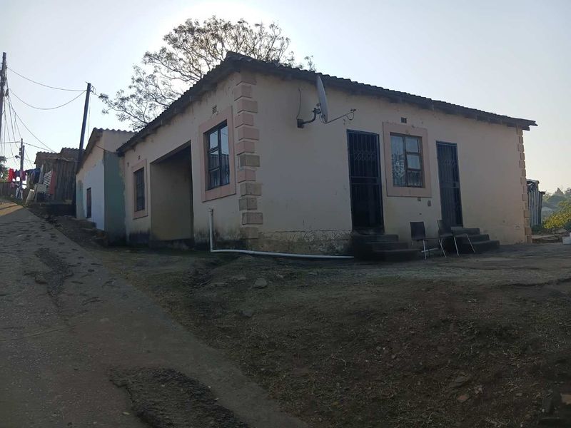 A single-story residential house featuring a tiled roof, security gates, and metal window frames, situated on an unpaved, uneven plot.