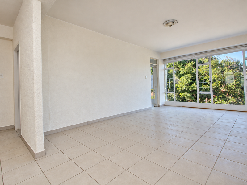A bright, empty living area featuring neutral tiled flooring, textured walls, and large windows that provide ample natural light and garden views.