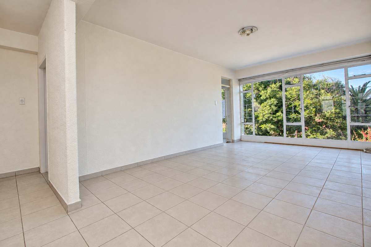 A bright, empty living area featuring neutral tiled flooring, textured walls, and large windows that provide ample natural light and garden views.