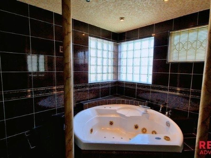 A corner spa bath featuring dark ceramic wall tiles, decorative columns, and glass block windows for natural light.