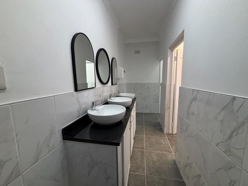 A clean, modern office bathroom featuring a triple vanity with vessel sinks, stone countertops, and marble-look wall tiling.