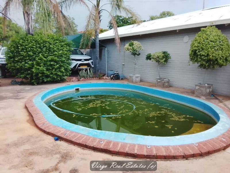 Outdoor pool area featuring a brick-edged swimming pool set against a painted brick exterior wall, currently requiring maintenance and cleaning.