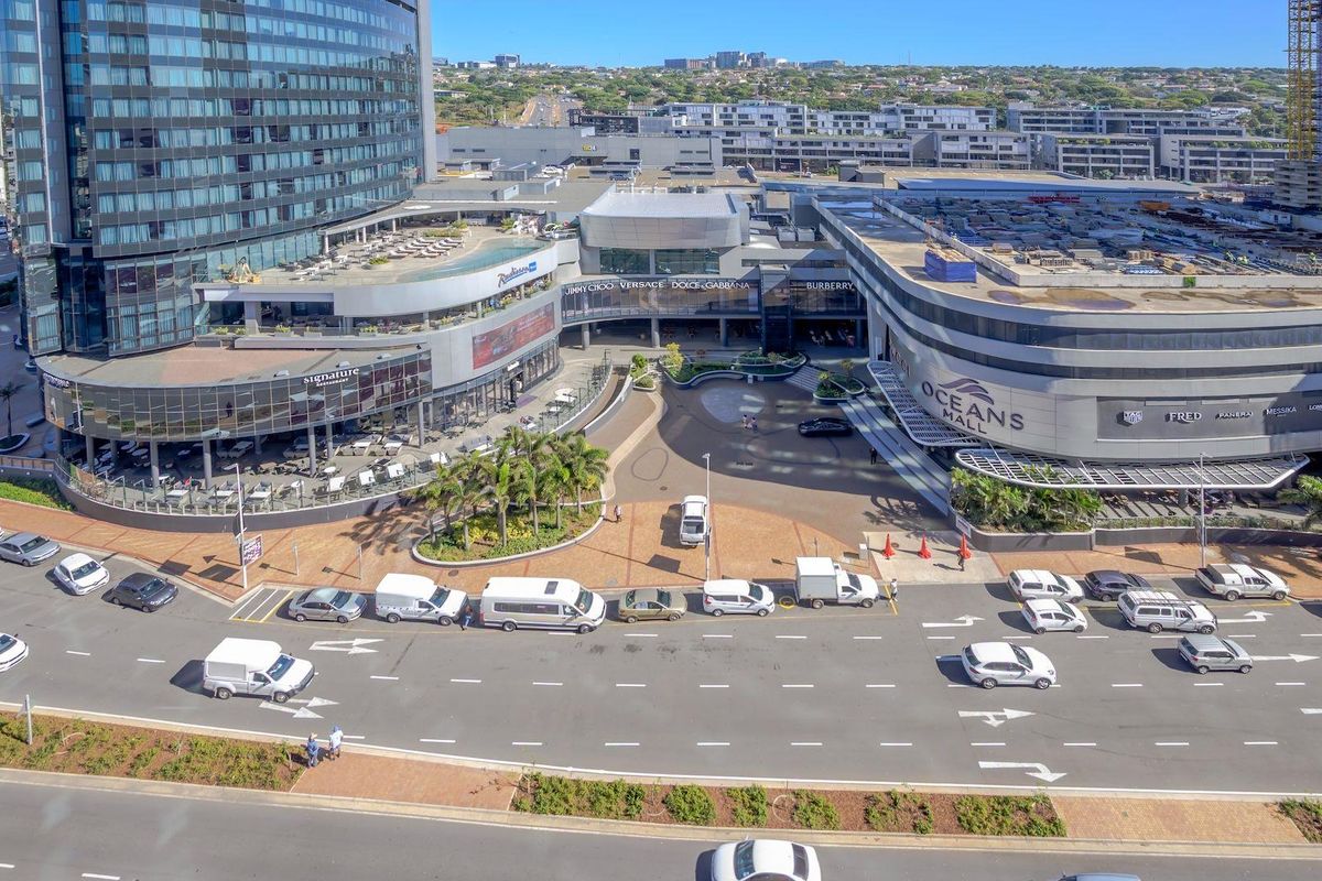 Aerial view of a modern commercial complex featuring a shopping mall, hotel tower, and paved plaza area with surrounding road infrastructure.