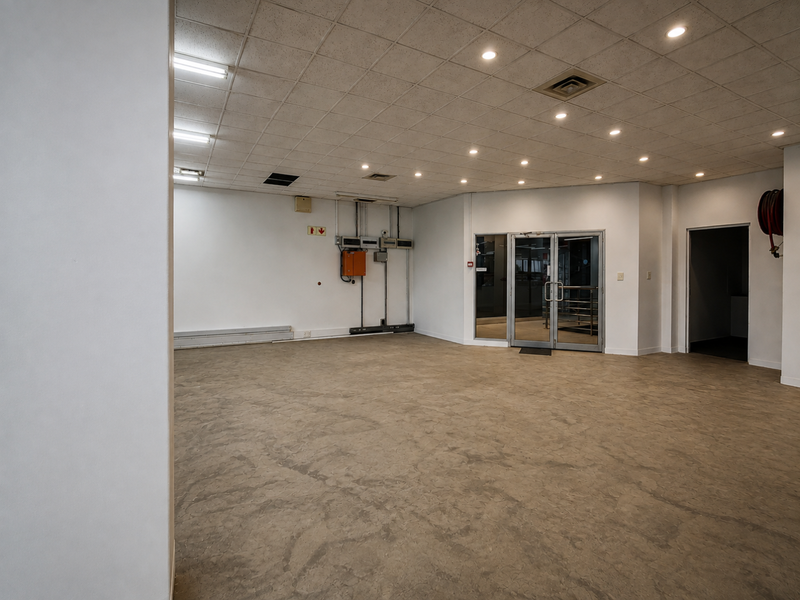 A spacious, empty office area featuring a suspended ceiling with recessed lighting, durable vinyl flooring, and a glass entrance door.