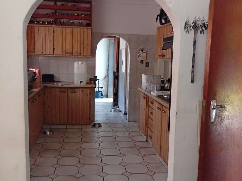 A dated kitchen featuring wooden cabinetry, tiled flooring, and a distinctive arched entryway leading to a secondary utility or scullery area.