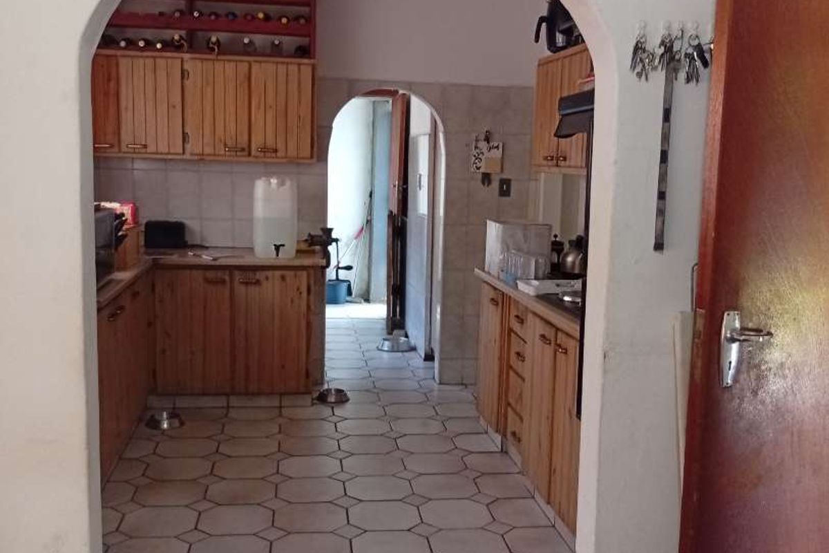 A dated kitchen featuring wooden cabinetry, tiled flooring, and a distinctive arched entryway leading to a secondary utility or scullery area.