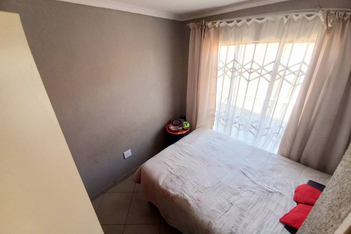 A compact bedroom featuring tiled flooring, neutral wall tones, and a window fitted with security bars and curtains.