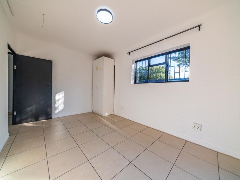 A bright, empty bedroom featuring tiled flooring, a built-in cupboard, and a window with security bars.