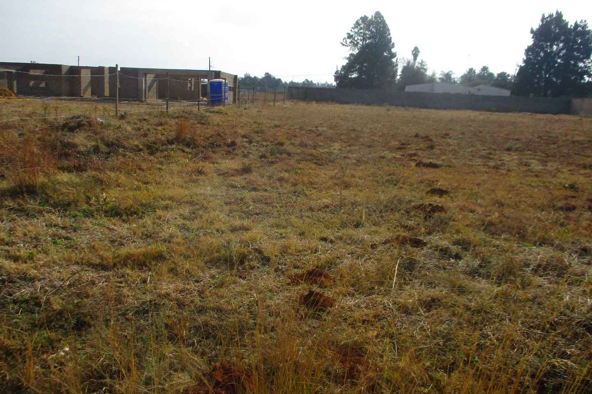 A view of a vacant residential plot featuring dry grass, perimeter fencing, and a neighboring structure under construction.