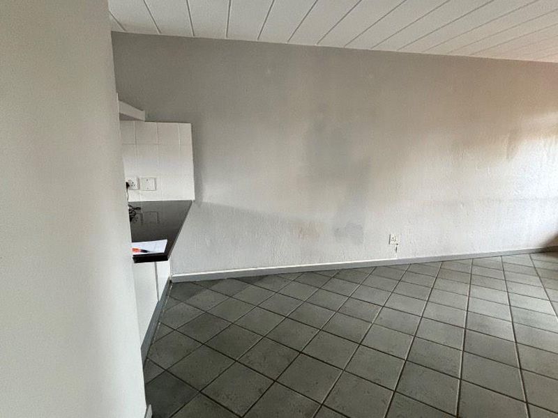 An empty open-plan living and kitchen area featuring tiled flooring, a panelled ceiling, and a compact kitchen corner with a tiled splashback.