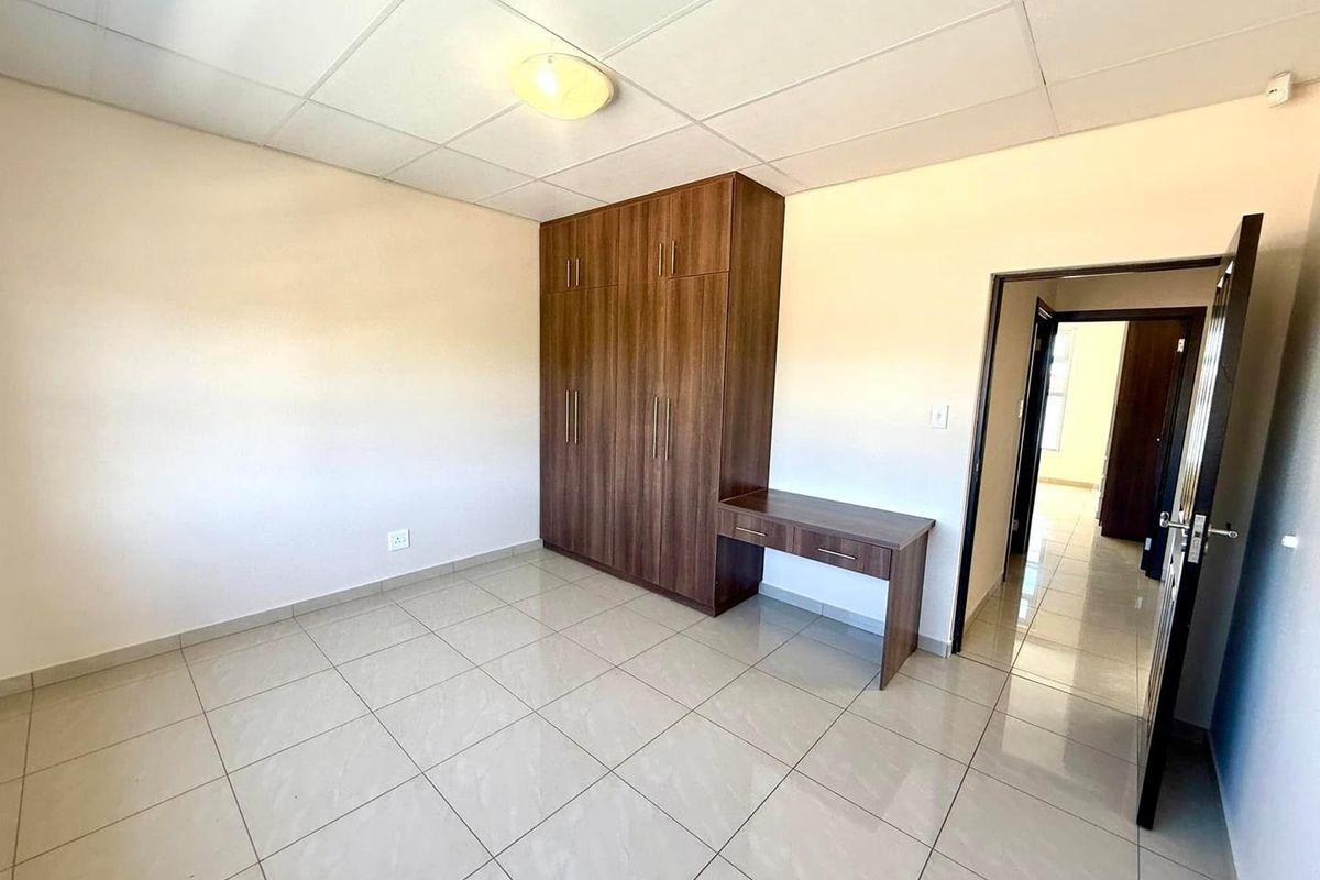 A bright, empty bedroom featuring neutral tiled flooring, a drop ceiling, and a modern built-in wooden wardrobe with an integrated desk unit.