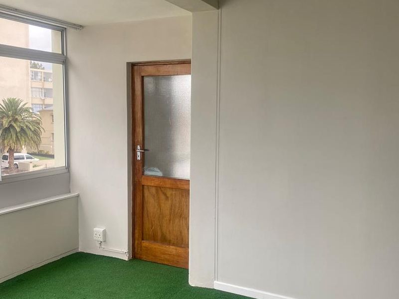 An enclosed balcony or sunroom featuring artificial grass flooring, a timber door, and large windows providing natural light.