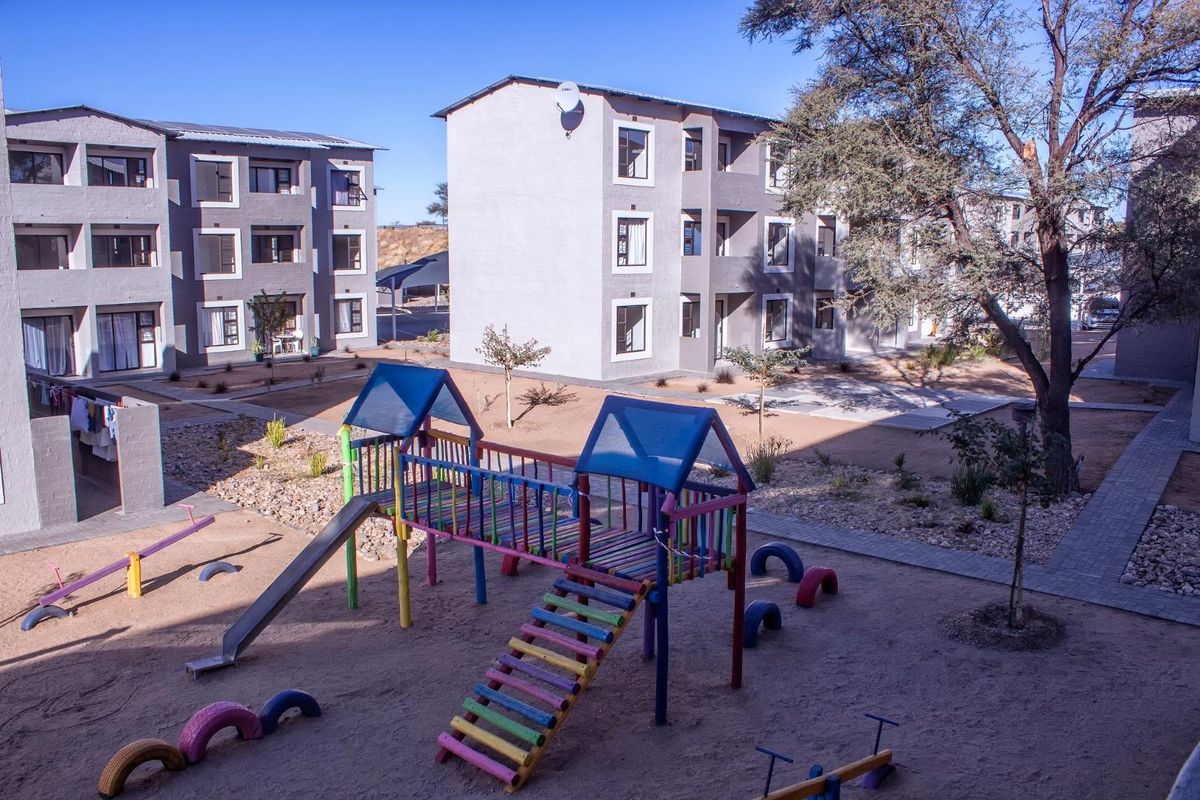 Exterior view of an apartment complex courtyard featuring a communal playground area with sand surfacing and paved walkways between residential buildings.