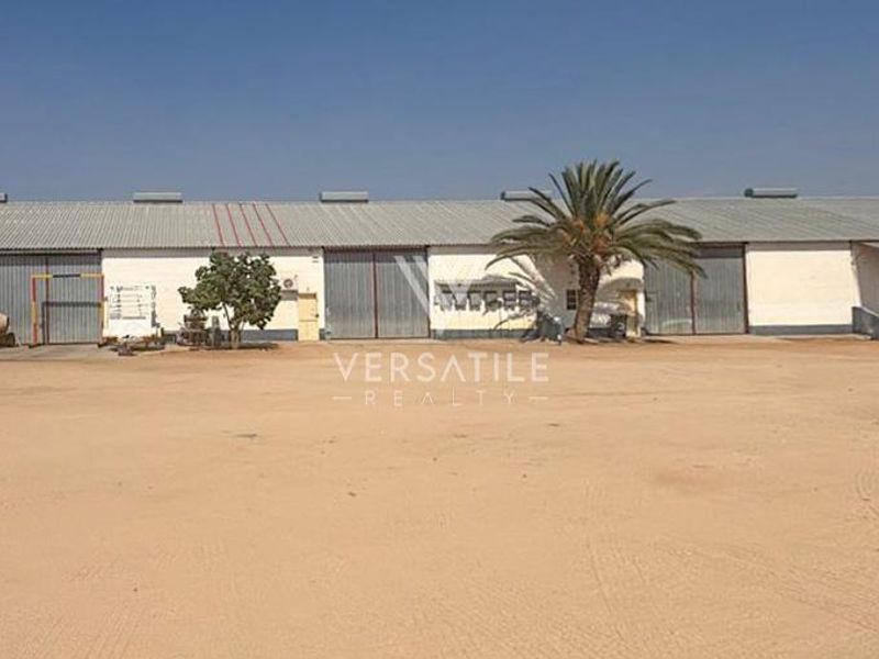 A wide-angle view of an industrial warehouse facility featuring large sliding doors, a corrugated metal roof, and a spacious unpaved yard area.