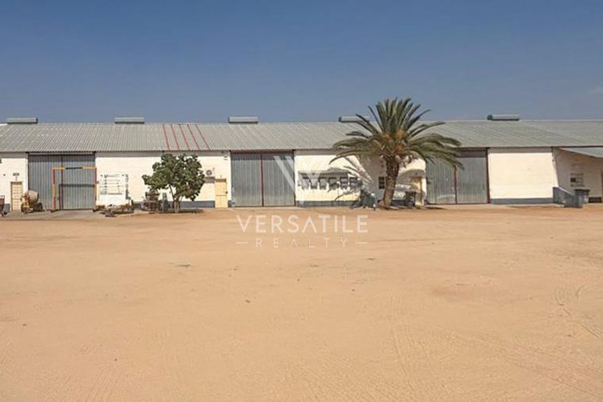 A wide-angle view of an industrial warehouse facility featuring large sliding doors, a corrugated metal roof, and a spacious unpaved yard area.