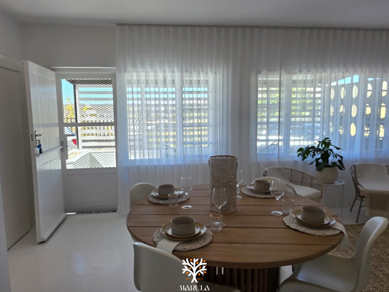 A bright dining area featuring tiled flooring, large windows with sheer curtains, and direct access to an exterior security gate.