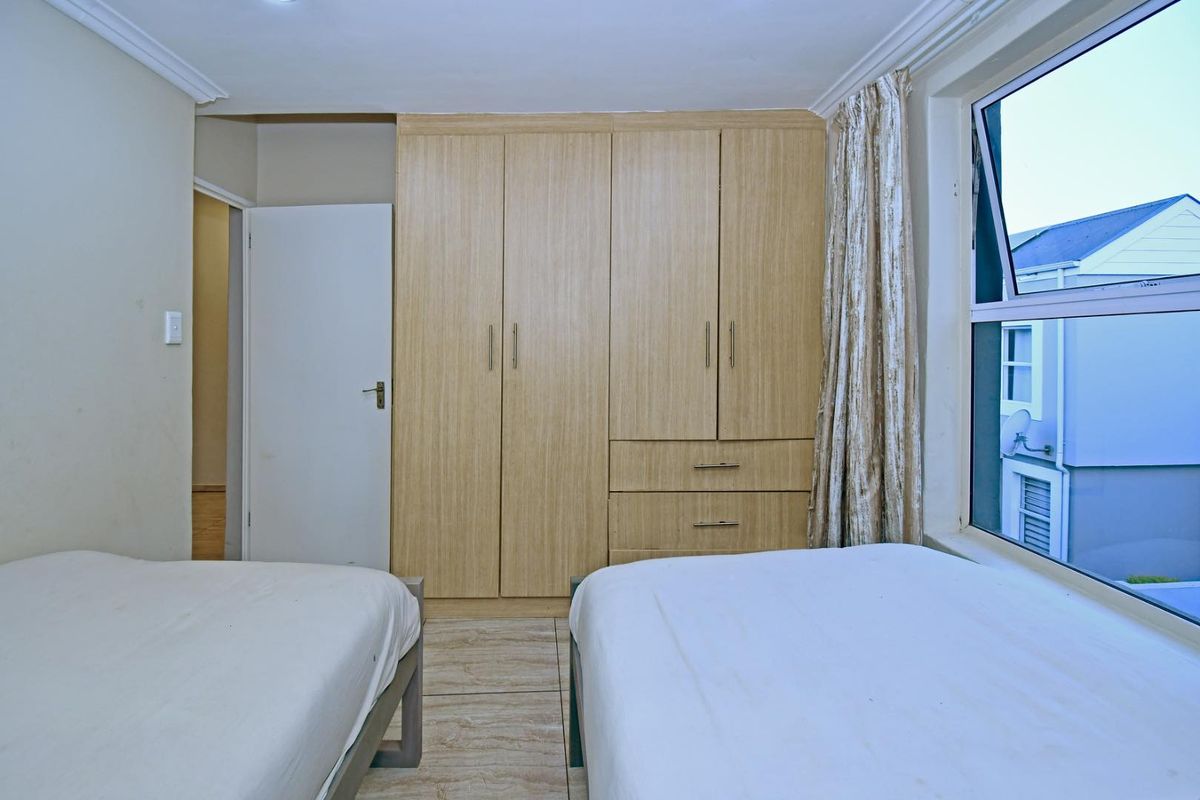 A functional bedroom featuring light wood-finish built-in cupboards, tiled flooring, and a large window providing natural light.