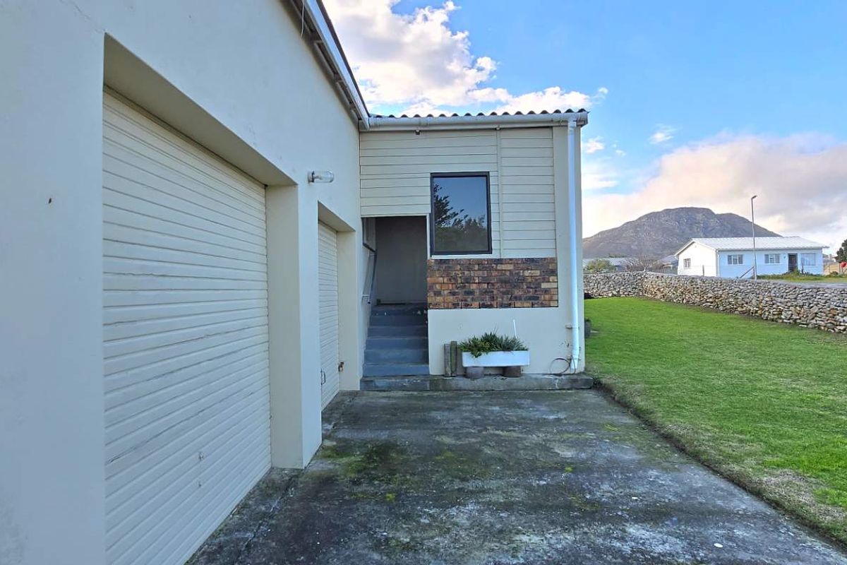 Exterior view of the property featuring a double garage with roller doors, a side entrance, and a spacious lawn area with stone boundary wall.