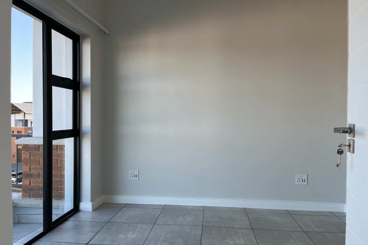 A bright, empty bedroom featuring neutral wall tones, tiled flooring, and a large black aluminium window providing natural light and access to a balcony.