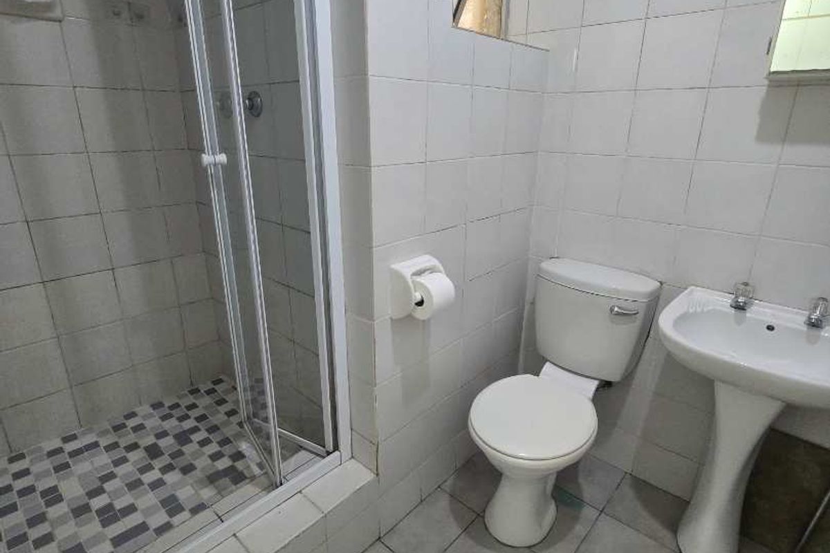 A functional bathroom featuring a shower enclosure, pedestal basin, and toilet, finished with white wall tiles and grey floor tiles.