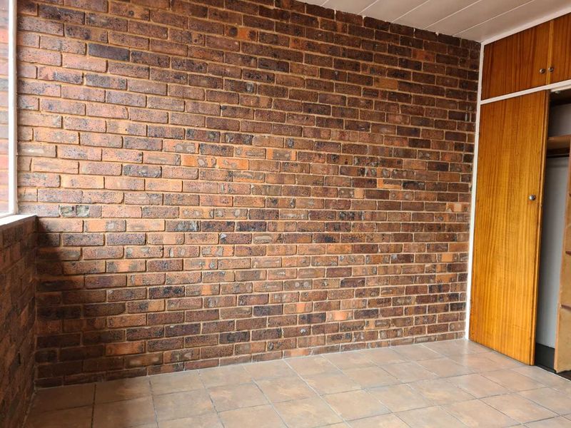 An empty bedroom featuring an exposed brick feature wall, tiled flooring, and a built-in wooden cupboard with overhead storage.