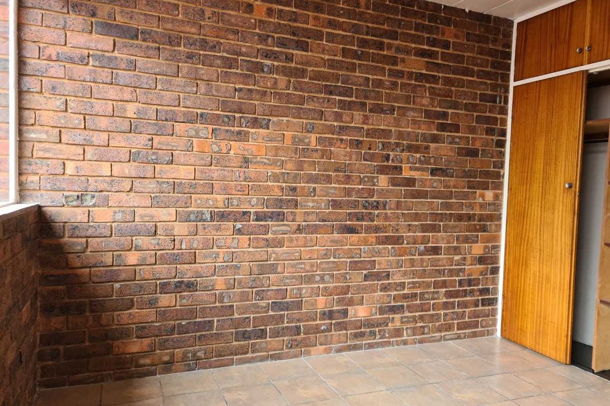 An empty bedroom featuring an exposed brick feature wall, tiled flooring, and a built-in wooden cupboard with overhead storage.