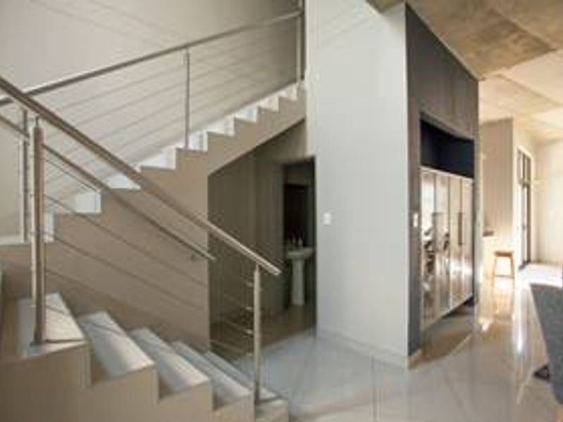 Modern hallway featuring a sleek stainless steel staircase railing, polished tiled flooring, and an exposed concrete ceiling, leading to an adjacent living area.
