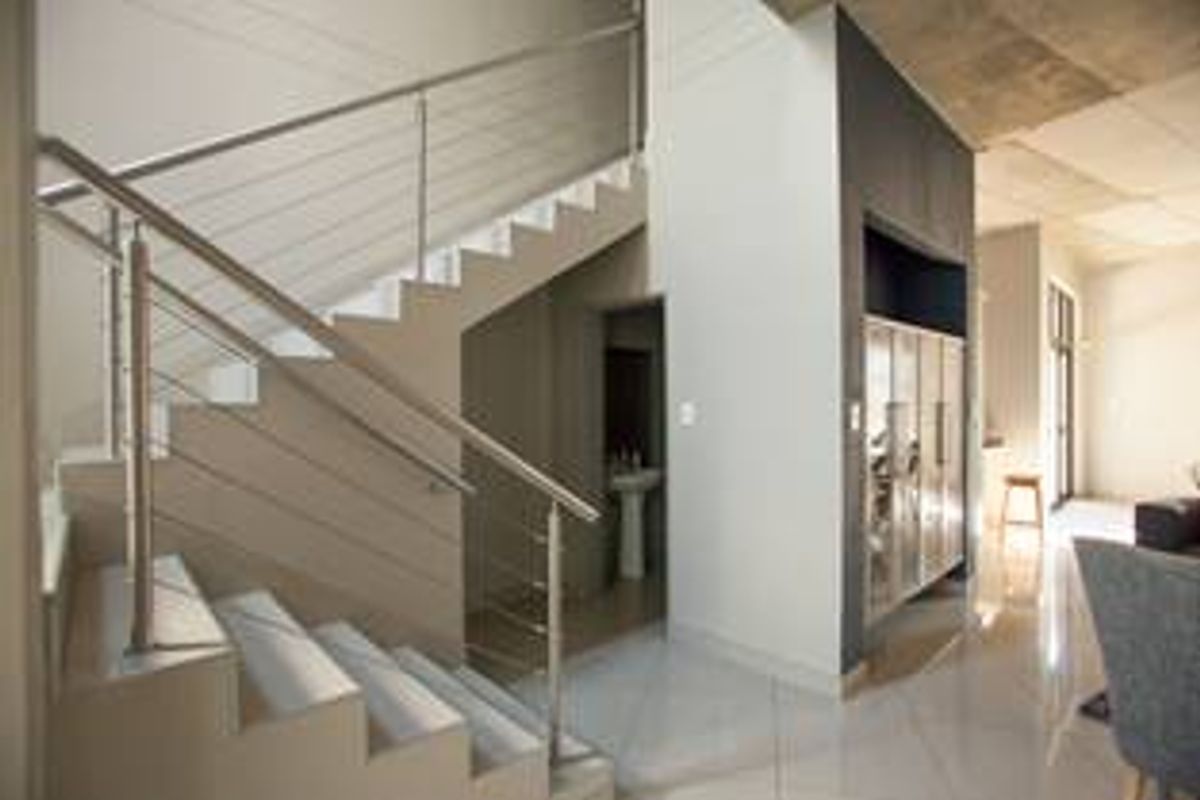 Modern hallway featuring a sleek stainless steel staircase railing, polished tiled flooring, and an exposed concrete ceiling, leading to an adjacent living area.
