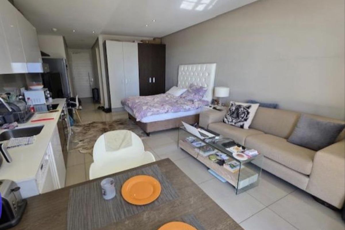 An open-plan studio apartment featuring a compact kitchen with built-in cabinetry, tiled flooring, and recessed lighting, seamlessly integrating living and sleeping areas.