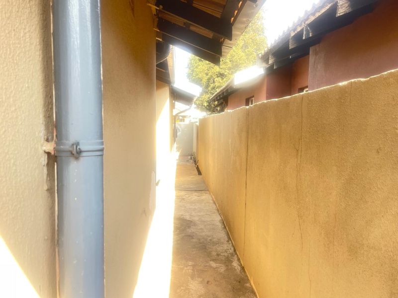 A narrow side passage between the house and a boundary wall, featuring a concrete path, downpipe, and exposed roof rafters.