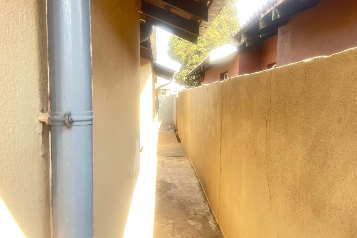 A narrow side passage between the house and a boundary wall, featuring a concrete path, downpipe, and exposed roof rafters.